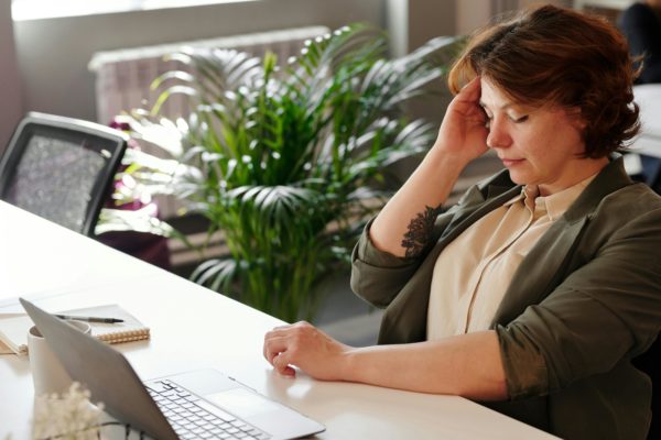 une femme assise à son bureau, fermant les yeux et se tient la tête, prise par la tristesse. 